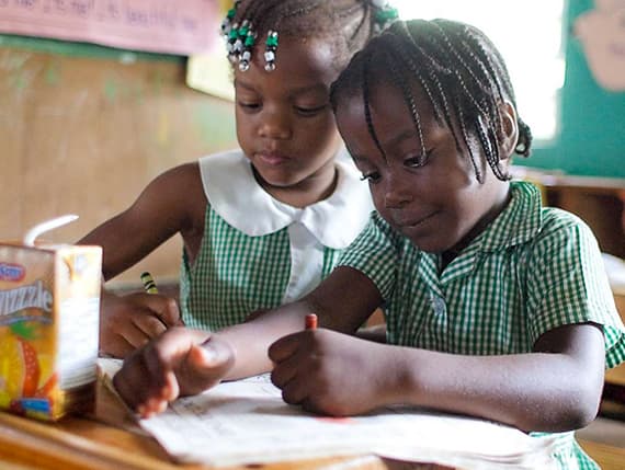 Two children in green checked uniforms writing at a shared desk at Little Bay All Age and Infant School